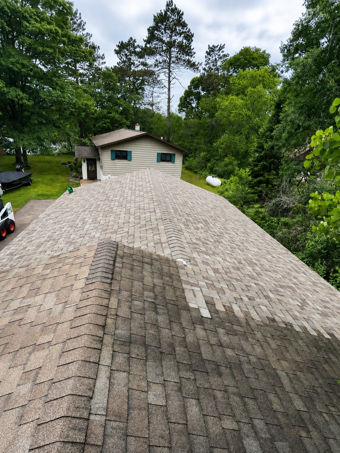 Completed roof of light driftwood-toned architectural shingles, photographed along the ridge of a wooded residential property